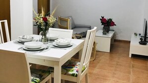 A dining area with a white table set for four, chairs, a small side table with a vase, and a television on a stand. - Impeccable 3-bed Apartment in Puerto de Sagunto (Sagunto)