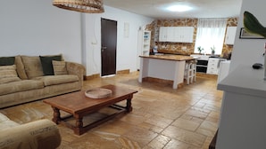 A living room with a sofa, a wooden coffee table, and a kitchen area with white cabinets and a tiled floor. - Andalusian Pool Parties, Music & Magic (Coin)