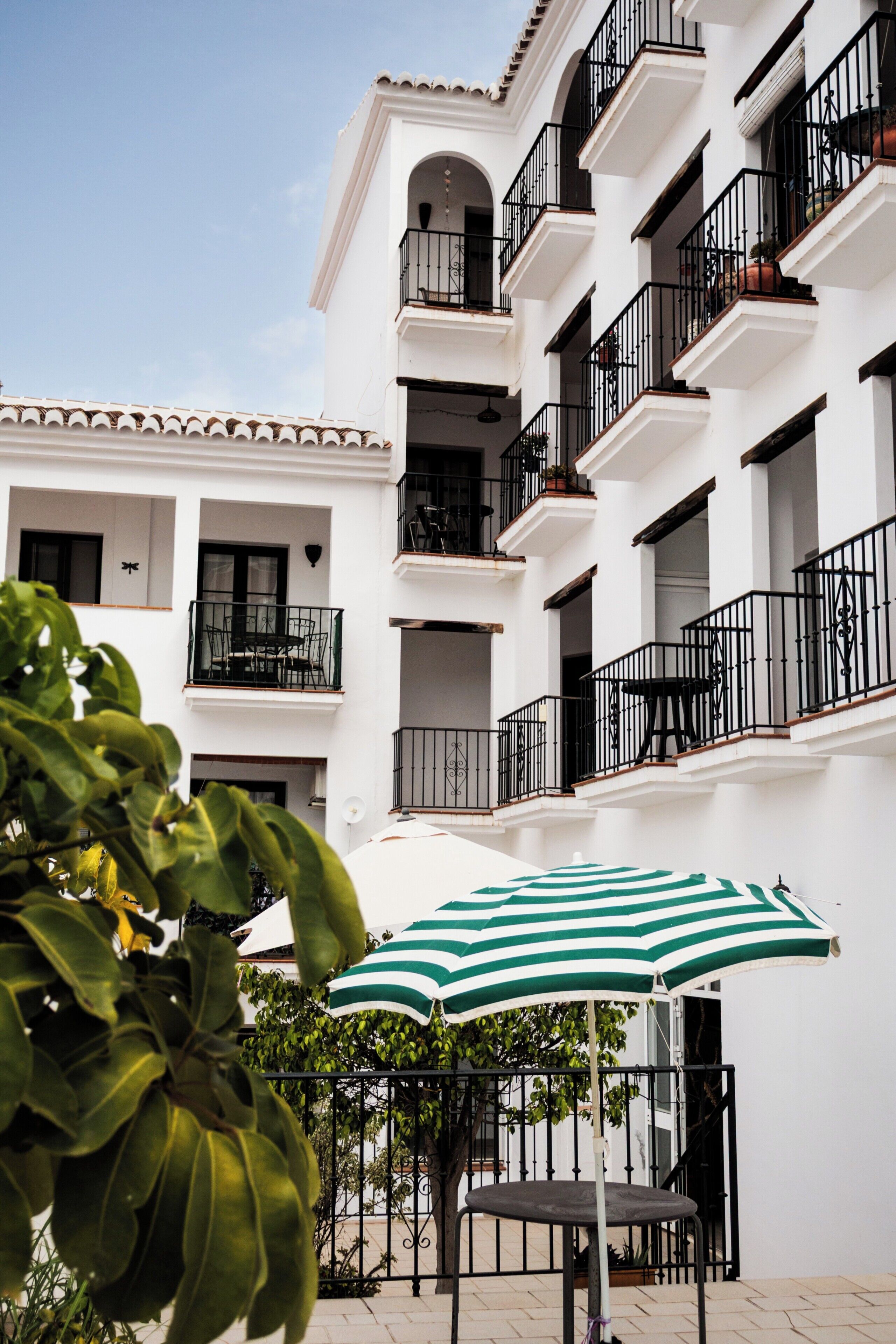 Un immeuble à plusieurs étages doté de balcons, un espace patio avec une table et des chaises, et un parasol à rayures vertes.