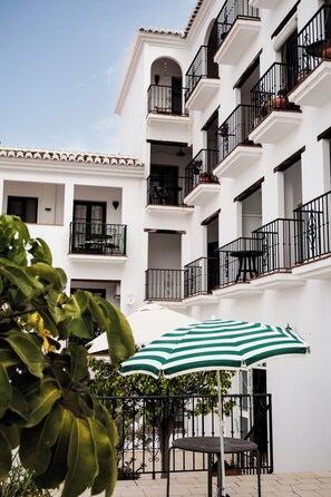 Un edificio de varios pisos con balcones, un patio con mesa y sillas, y una sombrilla a rayas verdes.