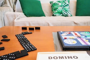 A table with dominoes and a board game, a sofa with green cushions, and a white wall in the background.