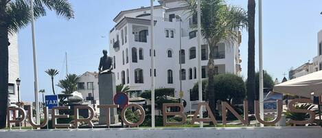 A large outdoor sign with the name "PUERTO BANUS" in bold letters, a statue of a man, and a building with balconies in the background.