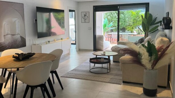 A modern living room with a dining area, a flat-screen TV mounted on the wall, and a large window offering a view of the outdoors. - Luxurious Apartment in Puerto Banus Marbella (Marbella)