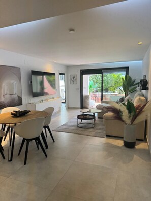 A modern living room with a dining area, a flat-screen TV mounted on the wall, and a large window offering a view of the outdoors. - Luxurious Apartment in Puerto Banus Marbella (Marbella)