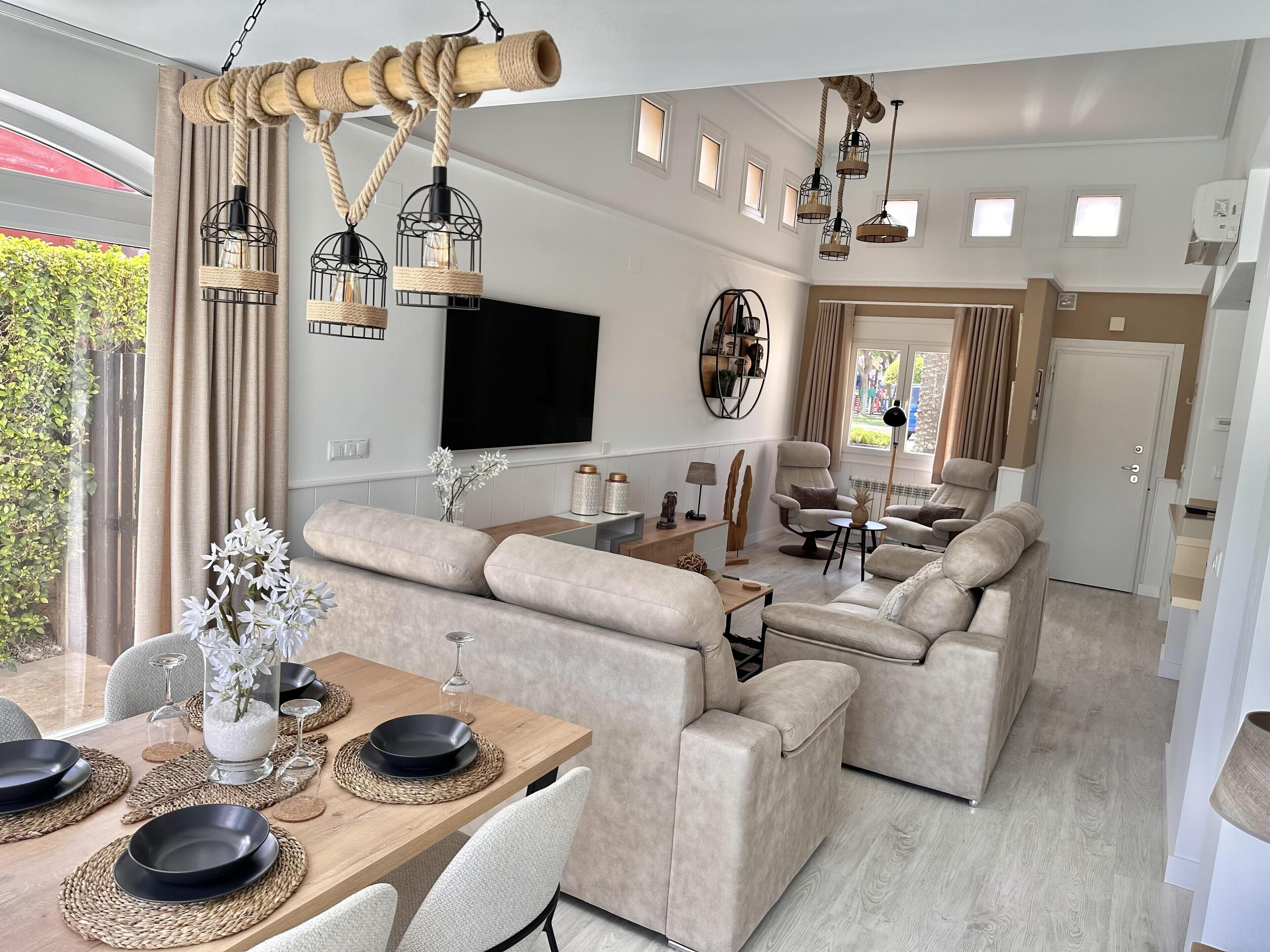 A modern living room with a large sectional sofa, a wooden dining table set for four, and a flat-screen TV mounted on the wall.