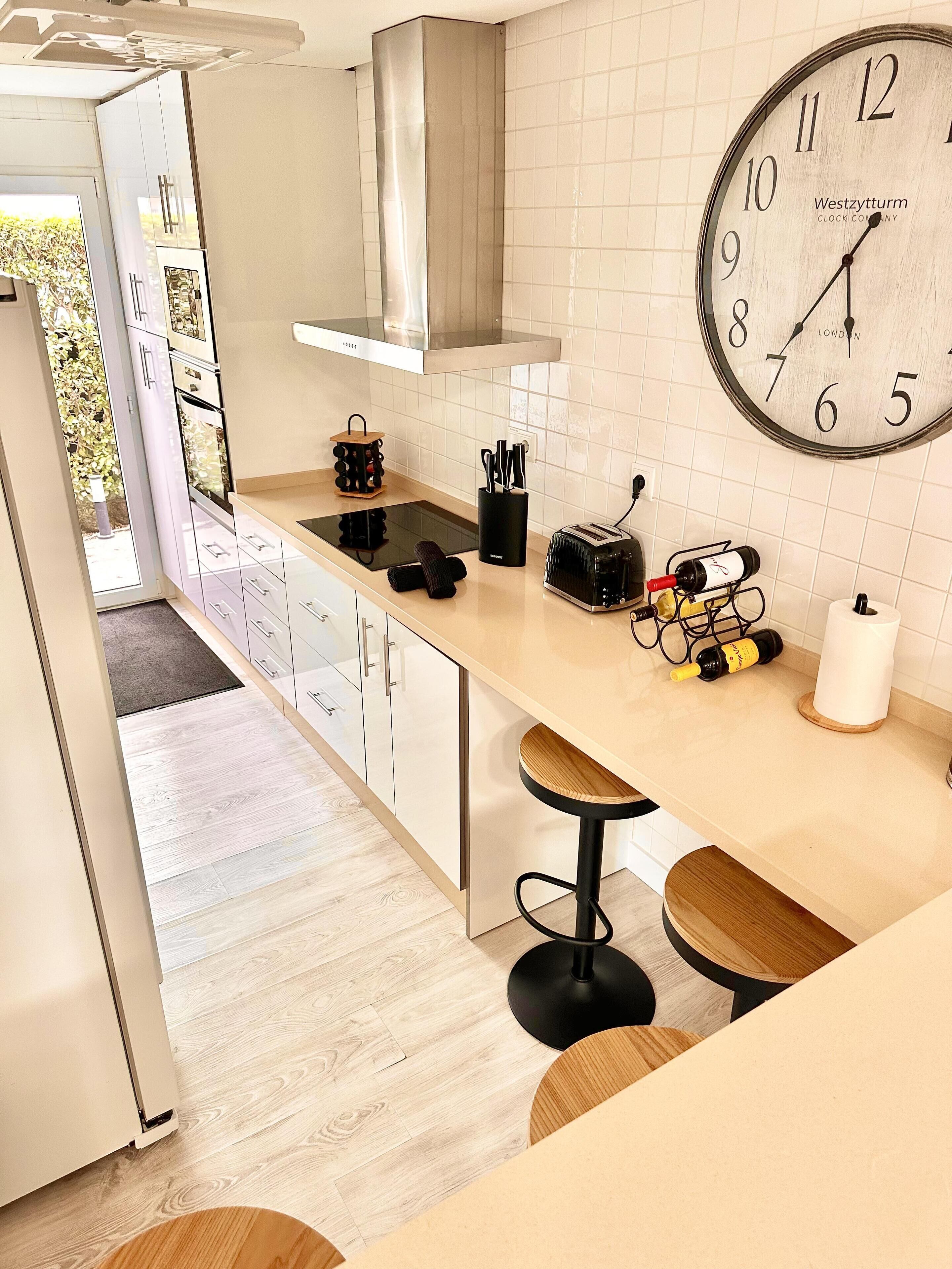 A modern kitchen with a Westzytturm clock, wooden bar stools, and a wine bottle on the counter.