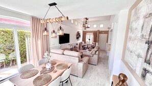 A modern living room with a sofa, dining table, and chairs. There is a TV mounted on the wall, and a large window with curtains. - Casa Bonita Sleeps 4 With Private Pool (Torre-Pacheco)