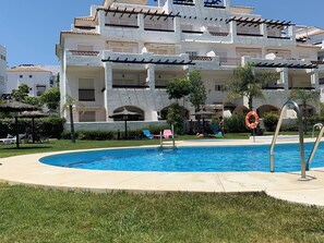 Une piscine entourée de chaises longues et de parasols, elle-même bordée d’une pelouse bien entretenue et d’un immeuble à plusieurs étages doté de balcons.