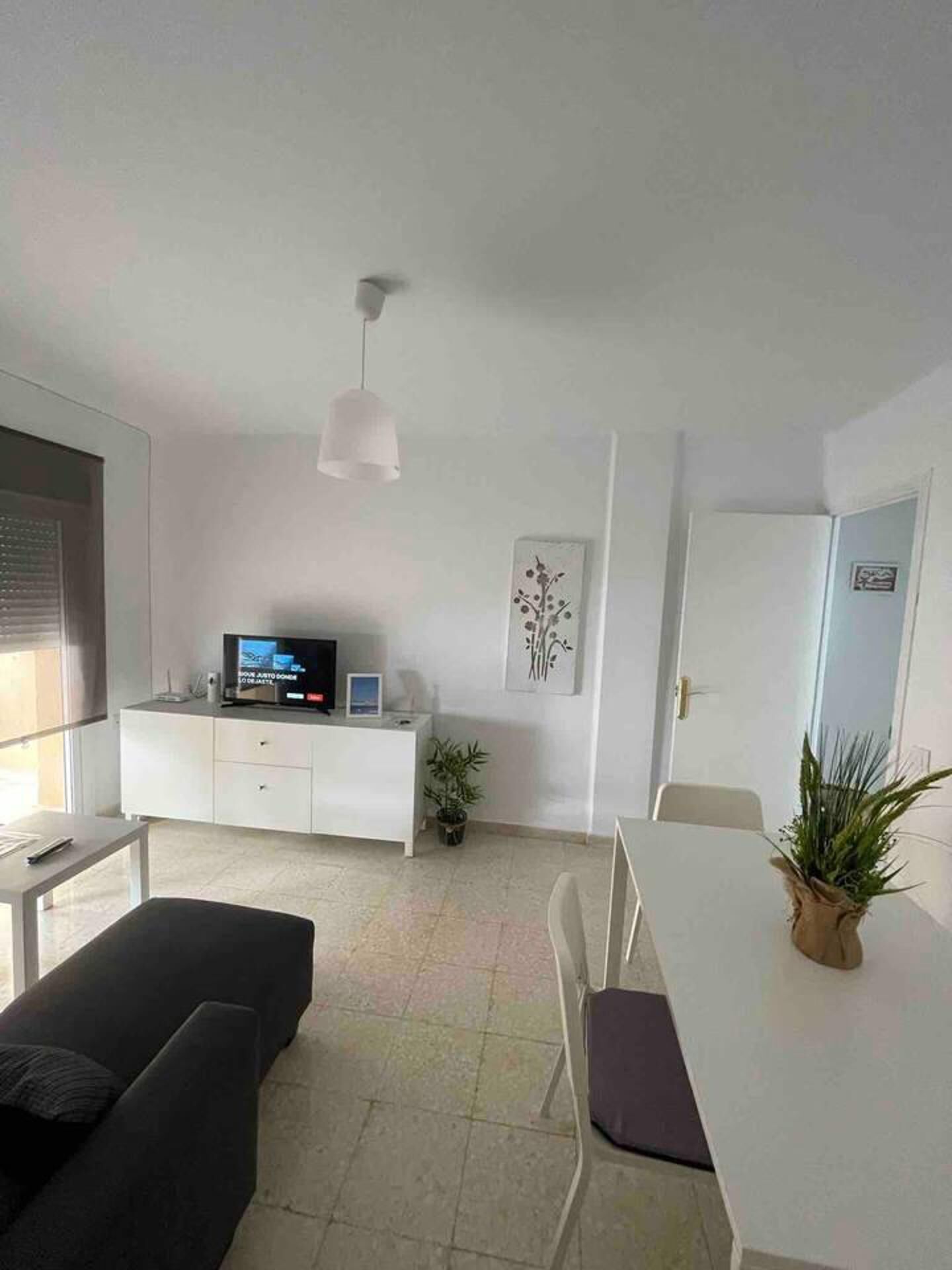 A modern, well-lit living room with a white sofa, a dining table, and a TV on a white cabinet.