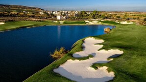 Egy golfpálya, amelyhez tartozik egy tó, homokakadályok, és a háttérben egy lakóövezet látható.
