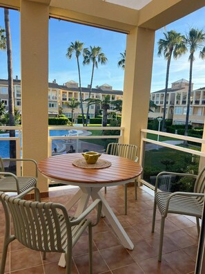 A balcony with a round table and chairs, overlooking a swimming pool and palm trees. - 2-bed Apartament Rosaleda Monterey (Denia)