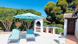 An outdoor seating area with two lounge chairs and a blue umbrella, surrounded by greenery and a stone patio. - Moraira Escape With Private Pool, 1 Bed, Sleeps 2 (Teulada)
