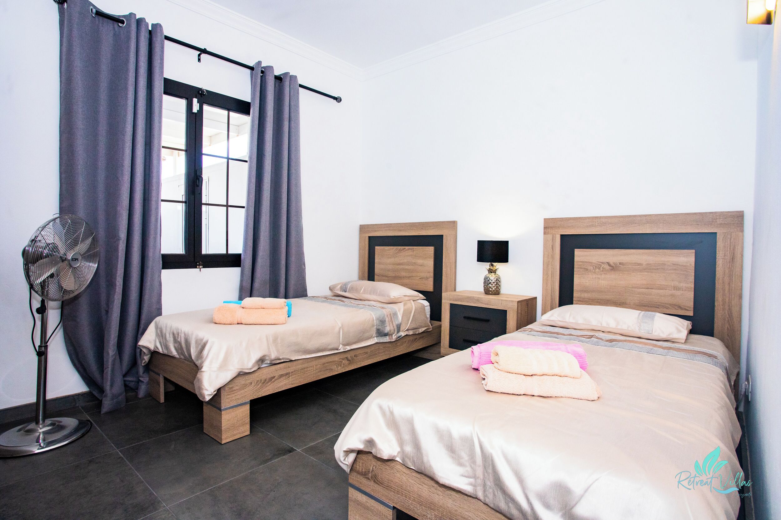 A hotel room with two wooden beds, a standing fan, a window with curtains, and a nightstand with a lamp.