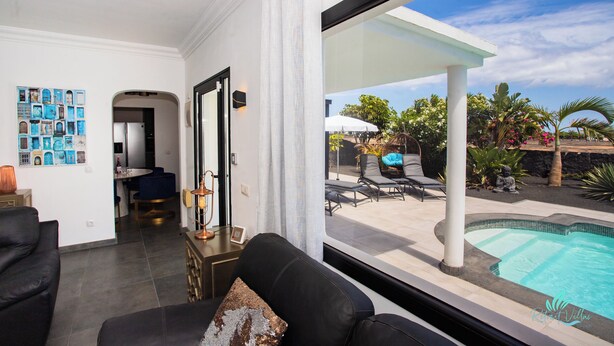 A modern living room with a pool view, featuring a black sofa, a dining area, and a wall with framed pictures. - Gorgeous Newly Renovated 4-bedroom Villa! (Yaiza)