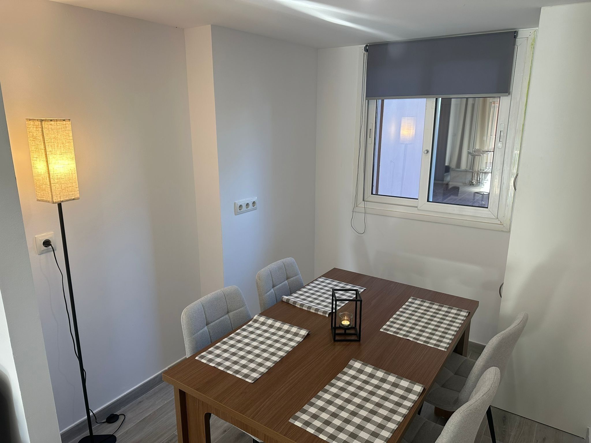 En matplats med ett träbord, fyra rutiga bordsunderlägg, två grå stolar, en golvlampa och ett fönster med gardiner.