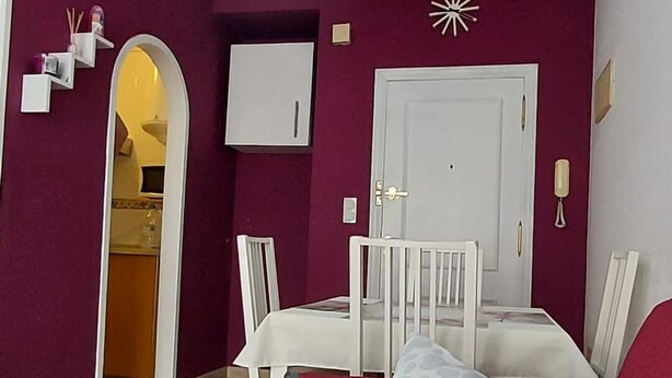 Egy piros szőnyeggel borított szoba, egy fehér és szürke mintás párnával ellátott ágy, egy fehér szék, egy kis asztal és egy fehér ajtó.