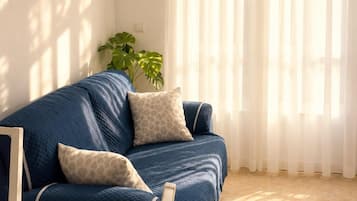 A room with a blue sofa, two pillows, a potted plant, a dining table, and a window with sheer curtains.