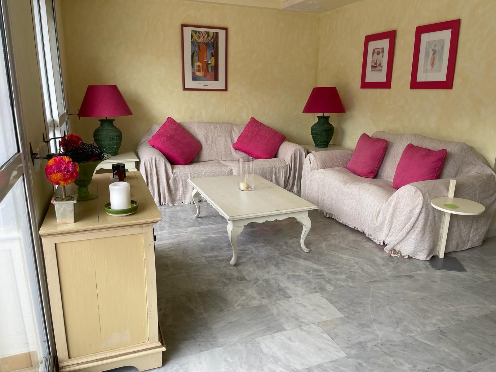 Un salon avec deux canapés, une table basse et deux lampes. Des tableaux encadrés ornent le mur, et un vase rempli de fleurs est posé sur une commode.