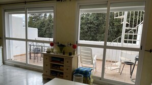 A room with large windows, a white table, a chair, a small cabinet, and a view of a balcony with outdoor furniture. - Beautiful 4 Bedroom Duplex Apartment (Estepona)