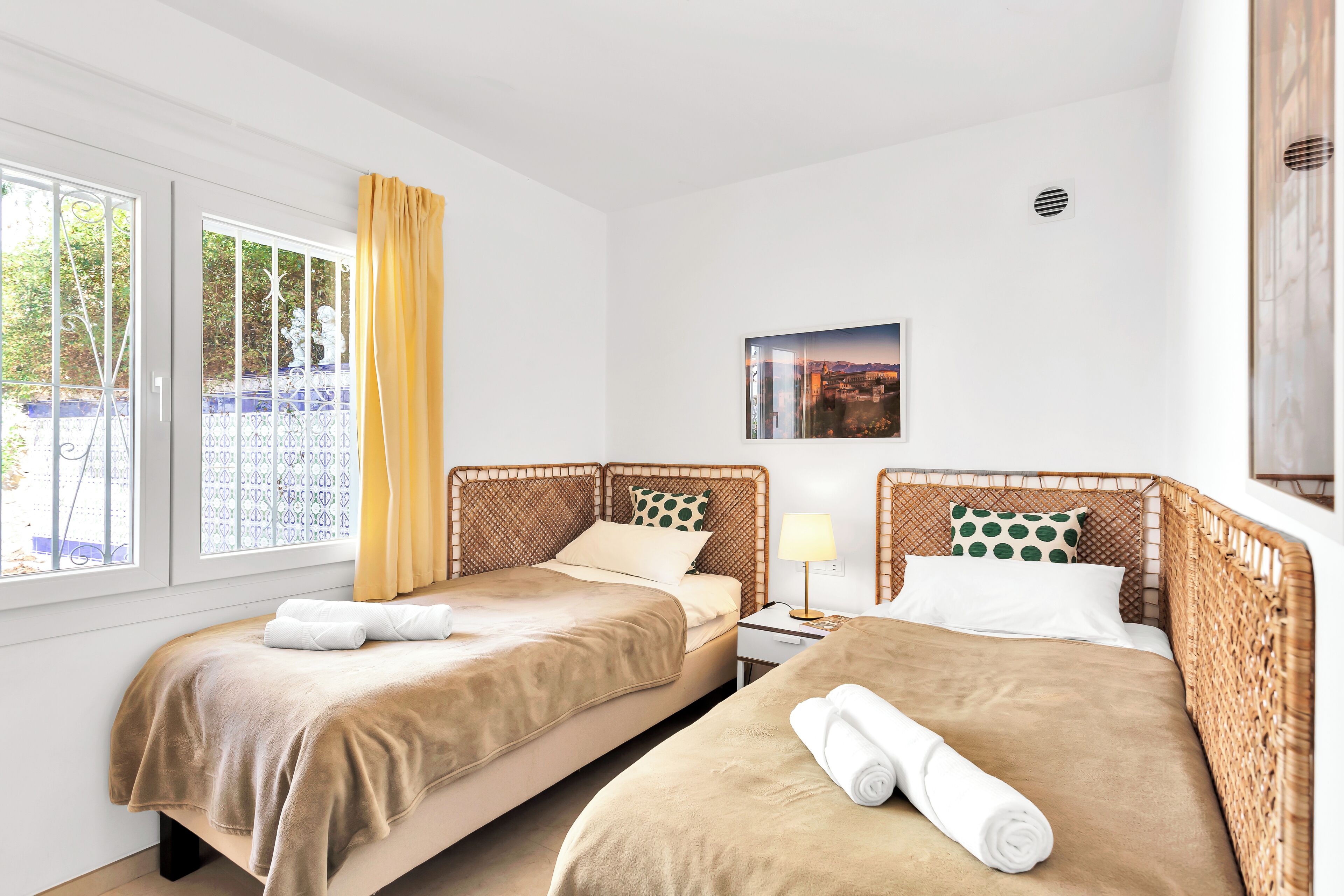 Habitación de hotel con dos camas, una ventana con cortinas, una mesita con lámpara y un cuadro en la pared.
