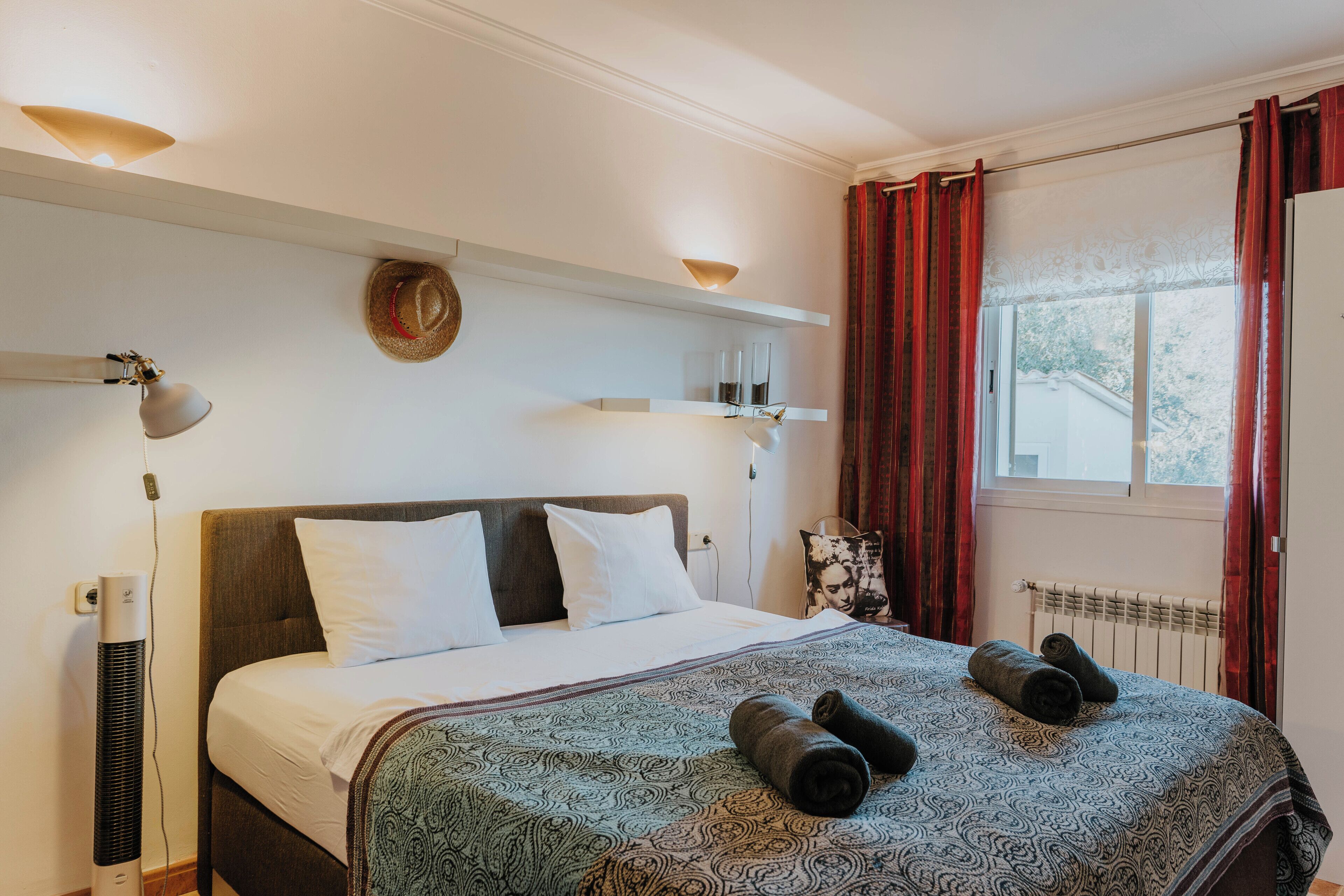 A neatly made bed with a patterned bedspread, a standing heater, and a window with curtains.