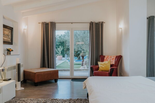 Une chambre à coucher comprenant un lit, un fauteuil rouge, un pouf marron, un radiateur et une porte-fenêtre coulissante donnant sur un jardin.