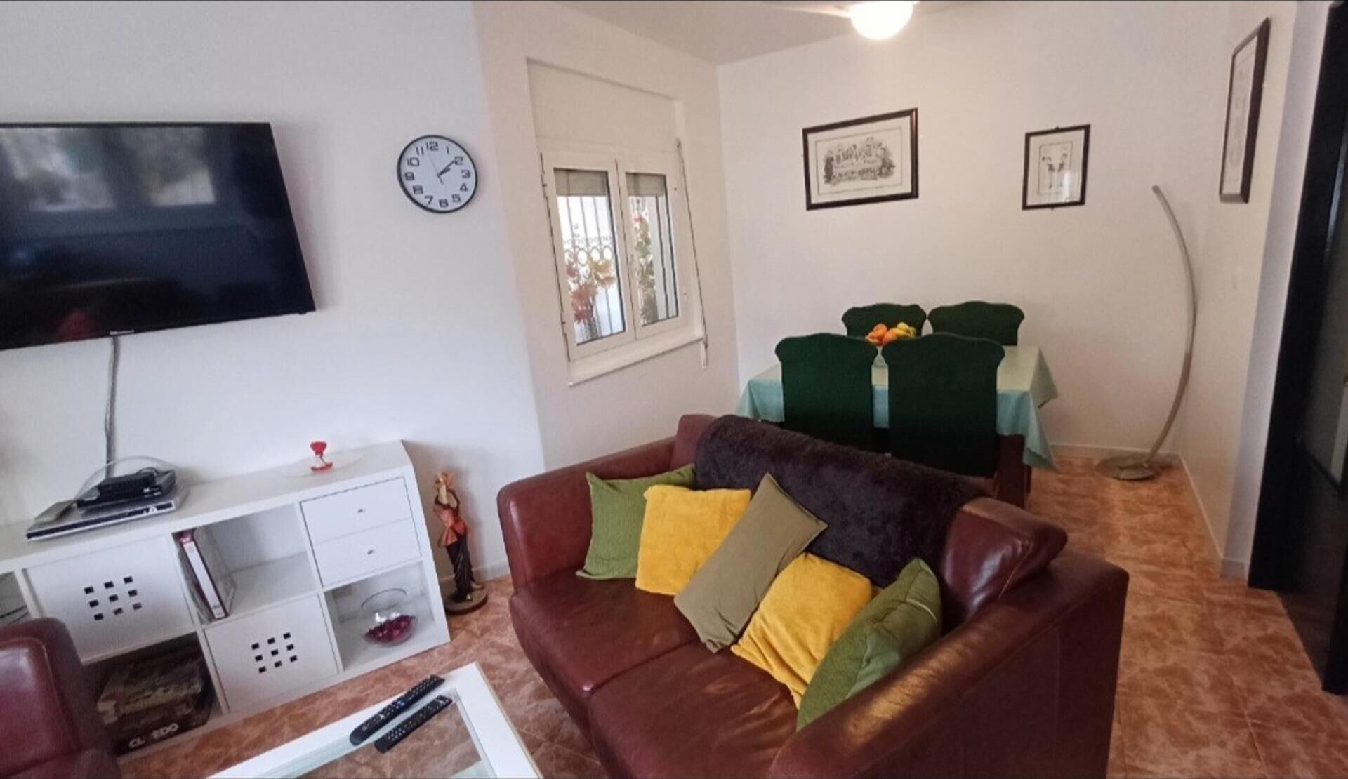 Een woonkamer met een bruine leren bank, een flatscreen televisie, een witte tv-kast, en een eethoek met groene stoelen.