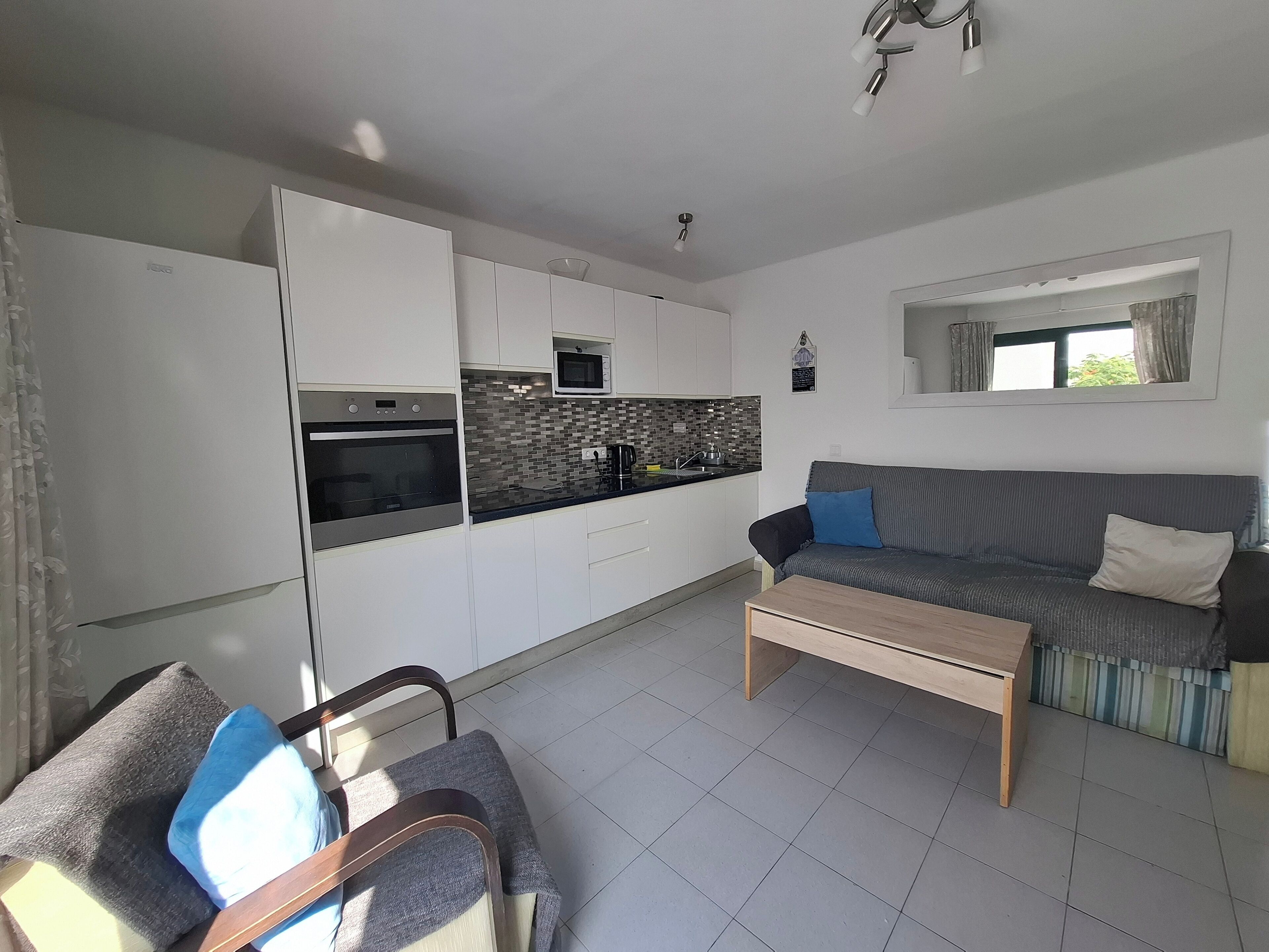 A modern kitchen with white cabinets, a black countertop, and a microwave. There is a grey sofa with blue and white cushions, a wooden coffee table, and a window with curtains.