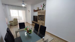 Un salon moderne avec une table à manger au plateau en verre, des chaises noires, un canapé beige, un téléviseur et un meuble en bois doté de portes en verre.