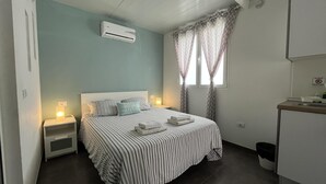 A bedroom with a bed, bedside tables, an air conditioner, a window with curtains, and a small kitchenette area. - Cosy Apartment in the Mountains Near Maspalomas (San Bartolomé de Tirajana)