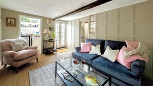 Una sala de estar con sofá, sillón y mesa de centro. Se ve una puerta que da a una cocina y una ventana que deja entrar luz natural.
