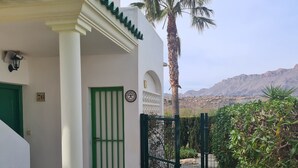 Un bâtiment à l’extérieur blanc, avec une porte verte et un hall d’entrée carrelé. On aperçoit un palmier et un portail au premier plan, et un paysage montagneux en arrière-plan.