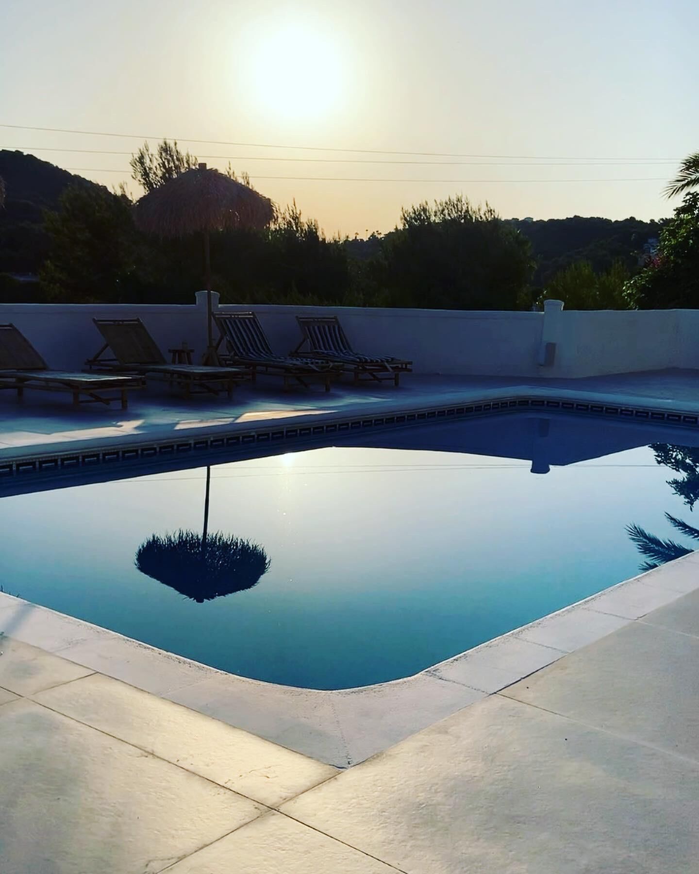 A swimming pool with a clear view of the sun setting in the background, surrounded by lounge chairs and a tiled deck.