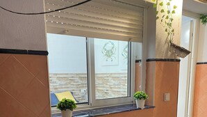 Un banco de madera con respaldo de listones, situado frente a una ventana que da a un muro de ladrillo y una puerta.