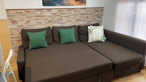 Un sofá marrón con cojines verdes y una almohada decorativa, frente a una pared con diseño de piedra y un cuadro de playa enmarcado encima.