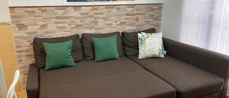 A brown sofa with green cushions and a decorative pillow, set against a stone-patterned wall with a framed beach painting above.