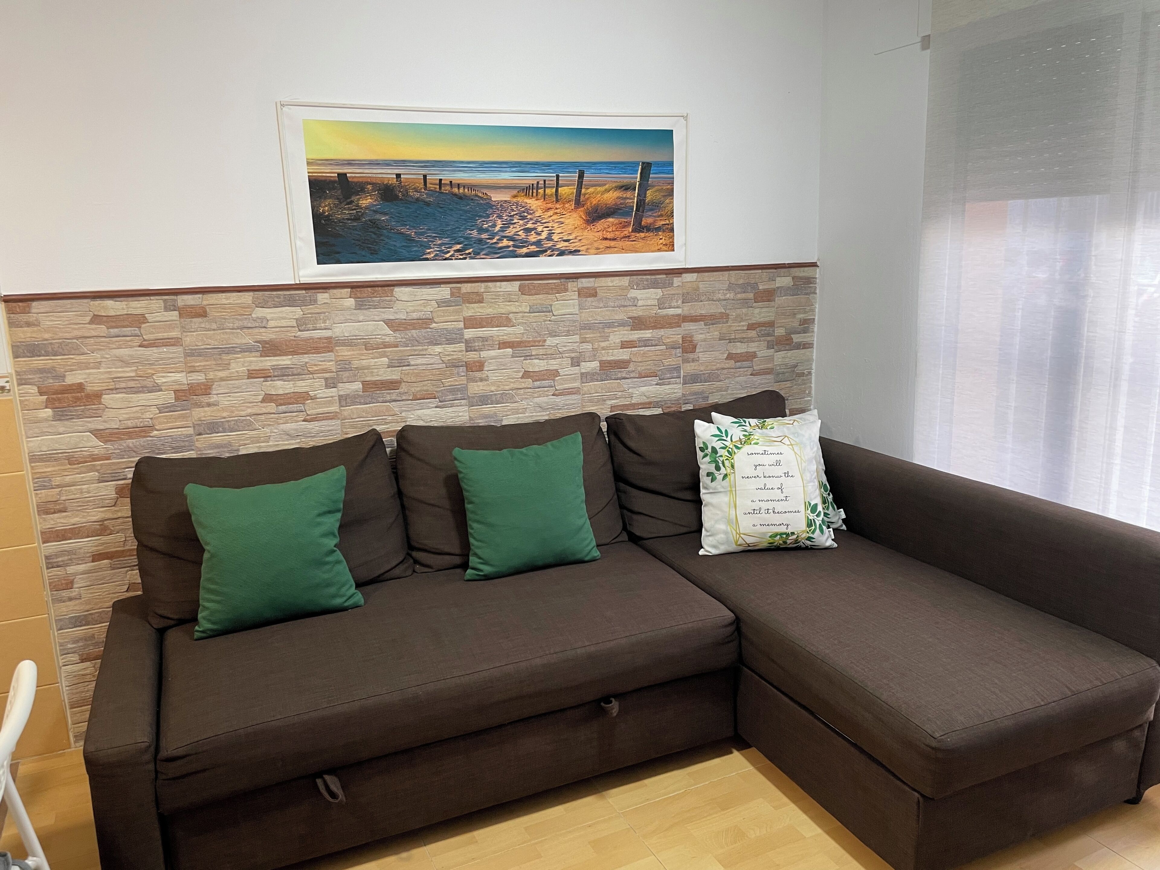 A brown sectional sofa with green cushions and a decorative pillow with a quote. A framed beach scene hangs above the sofa.