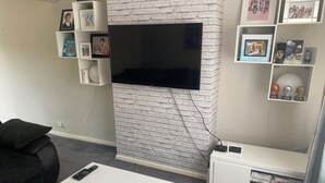 A living room with a flat-screen TV mounted on a white brick wall, a white coffee table with a remote control, a bookshelf with framed photos, and a white entertainment unit with a DVD player. - A real homely home close to Gatwick and the South East of England. (Crawley)