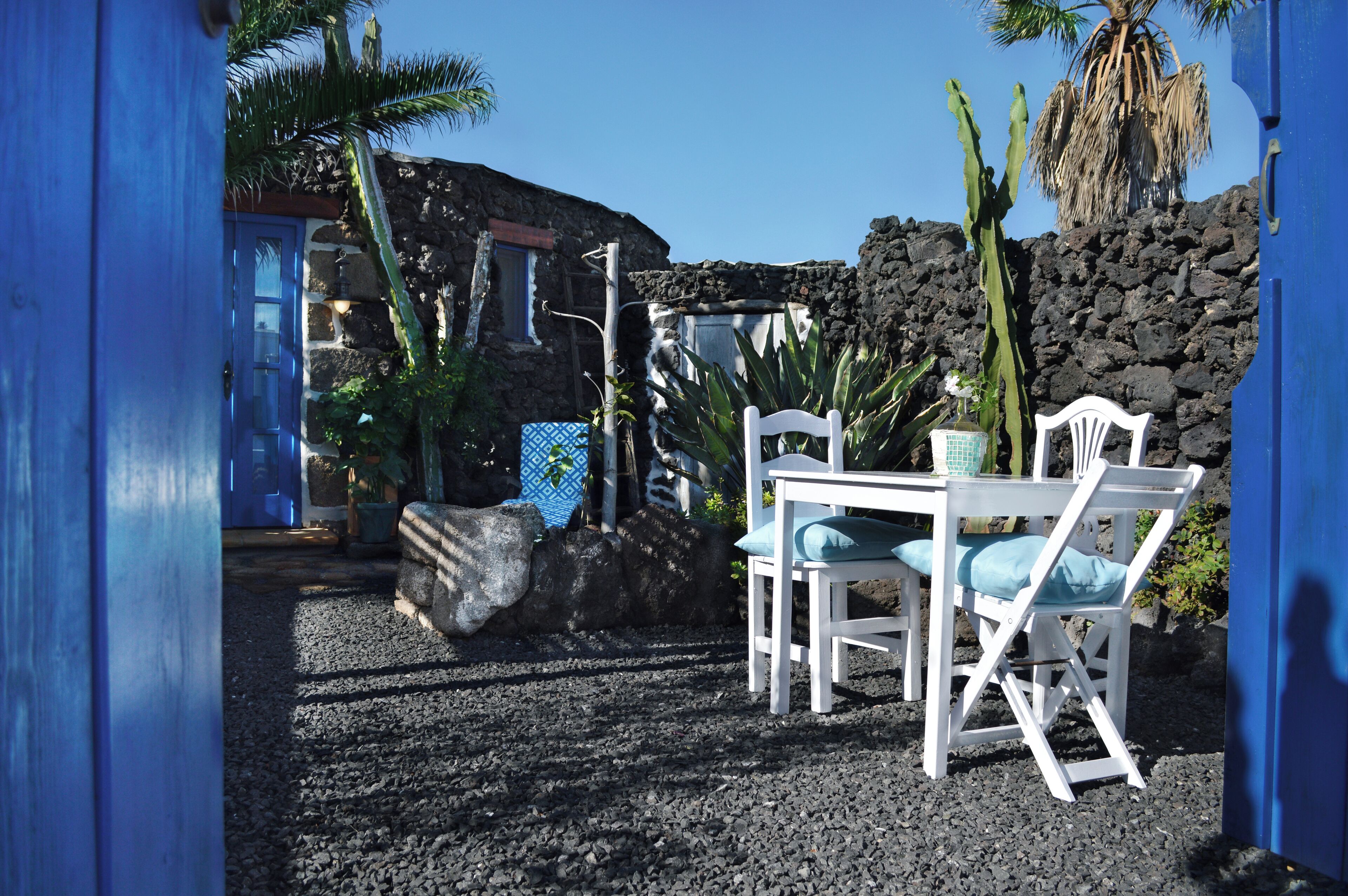 Un bâtiment en pierre avec une porte bleue, une table et des chaises blanches, et un cactus.