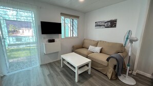 A living room with a beige sofa, a white coffee table, a flat-screen TV mounted on the wall, a standing fan, and a window with a view of the outside. - Renovated Tiny House in the Country Near Fataga (San Bartolomé de Tirajana)