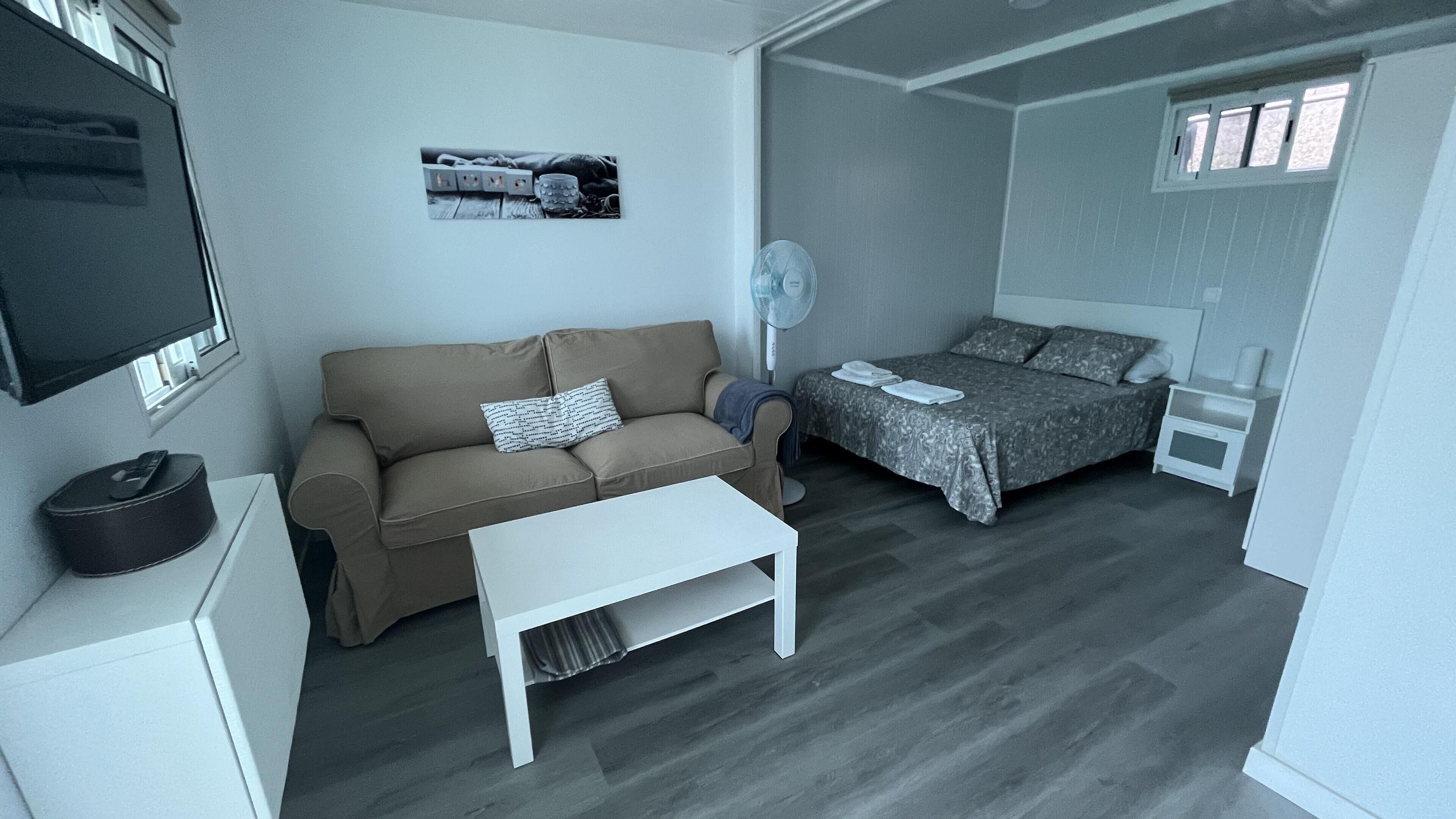 A compact room with a sofa, a small table, a bed with a patterned bedspread, a bedside table, a wall-mounted TV, and a framed picture.