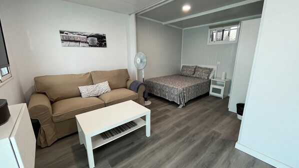 Une pièce de taille modeste comprenant un canapé, une petite table, un lit avec une couvre-lit à motifs, un ventilateur sur pied et une fenêtre avec des stores.