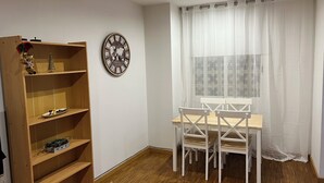 Ein Zimmer mit einem Bücherregal aus Holz, einer Wanduhr, einem Esstisch mit Stühlen und einem Fenster mit durchsichtigen Vorhängen.