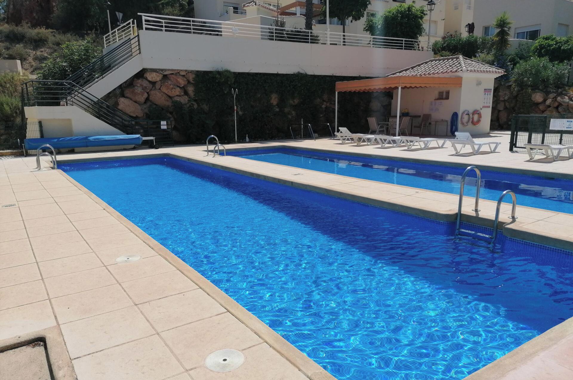 Une piscine rectangulaire à l’eau d’un bleu limpide, entourée d’une terrasse carrelée. On y trouve des chaises longues et un petit espace salon couvert. Un escalier mène à un niveau supérieur.