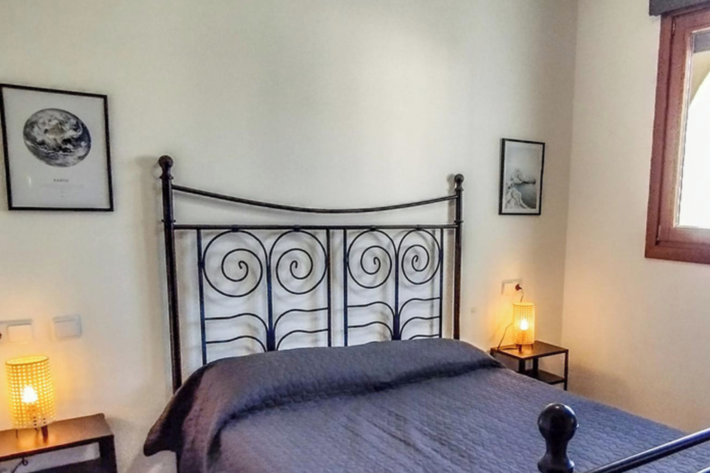 Un dormitorio con cabecera de metal, una cama con manta gris, dos mesitas de noche con lámparas y cuadros enmarcados en las paredes.