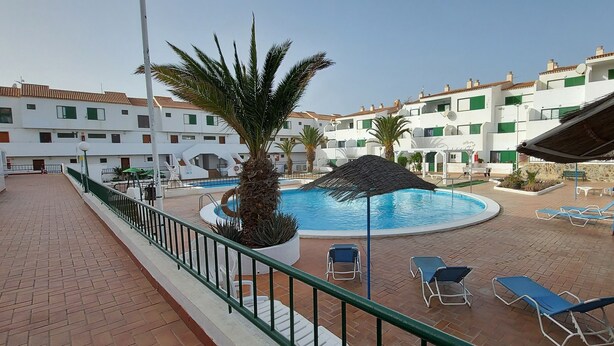 A pool area with lounge chairs, a palm tree, and a building with balconies in the background. - 1 Bed Apartment - Sleeps 2 - Pool - Parking (Arona)