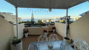 Eine überdachte Terrasse im Freien mit Tisch, Stühlen, Blumentöpfen und Blick auf das Meer.