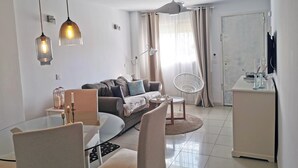 Ein modernes Wohnzimmer mit einer Couch, einem Glastisch und einem weißen Sessel. Ein Deckenventilator und Pendelleuchten sind vorhanden.