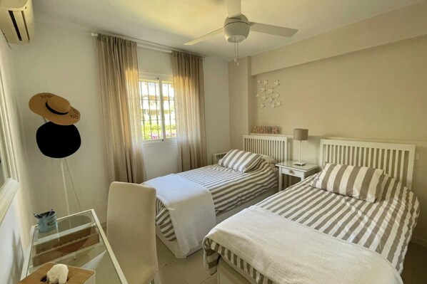 A hotel room with two beds, a ceiling fan, a window with curtains, a radiator, and a small table with a lamp.