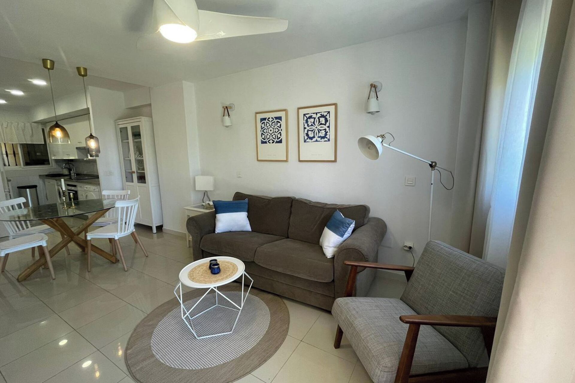 Una sala de estar moderna con sofá, sillón, mesa de comedor y sillas. Se observa una cocina al fondo.